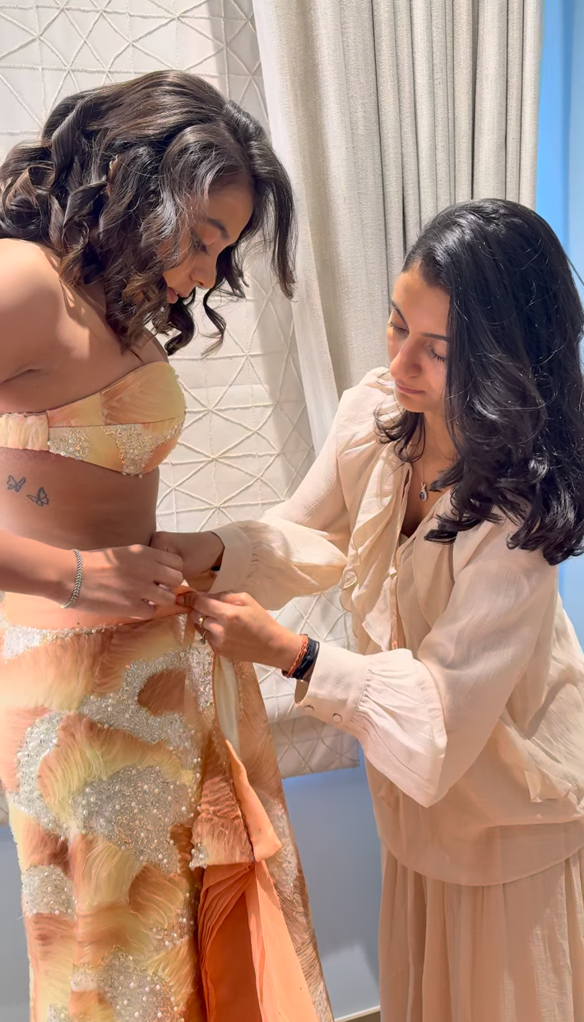 A woman in a peach and orange embellished saree has her hair styled by another woman in a cream-colored blouse and skirt set, set against an indoor backdrop with curtains.